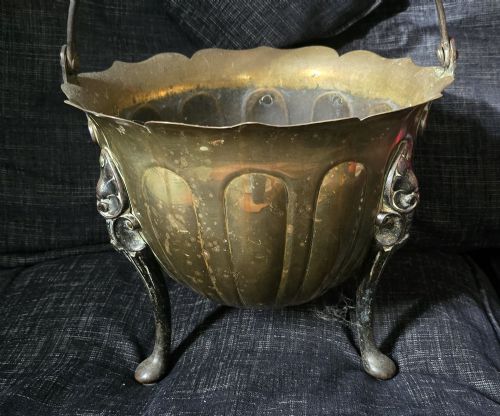 fig 1. An Arts and Crafts brass coal scuttle featuring a scalloped rim, vertical body fluting, three dark cast legs, and an overhead coiled handle.