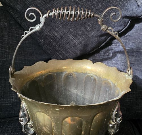 fig 2. An Arts and Crafts brass coal scuttle featuring a scalloped rim, vertical body fluting, three dark cast legs, and an overhead coiled handle.