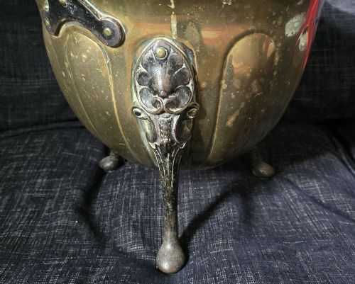 fig 3. An Arts and Crafts brass coal scuttle featuring a scalloped rim, vertical body fluting, three dark cast legs, and an overhead coiled handle.