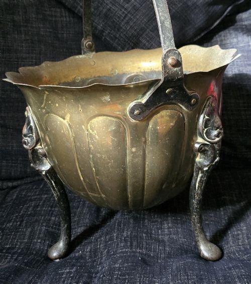 fig 9. An Arts and Crafts brass coal scuttle featuring a scalloped rim, vertical body fluting, three dark cast legs, and an overhead coiled handle.