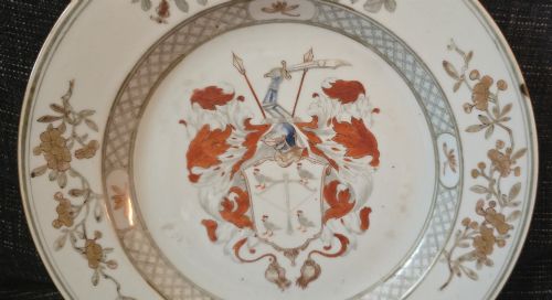 fig 2. A circular Chinese export porcelain plate featuring a central heraldic crest with red mantling, encircled by a geometric diaper border and floral sprigs.