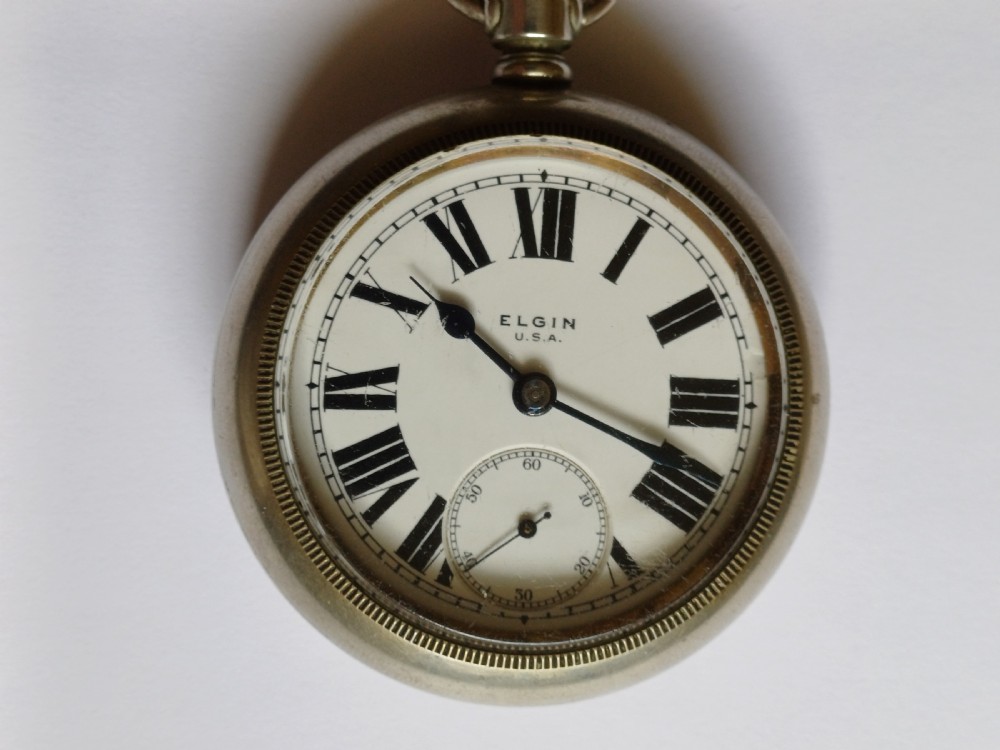 An open-face antique pocket watch featuring a white enamel dial with black Roman numerals, a sub-seconds dial, and a silver-tone nickel case. View III