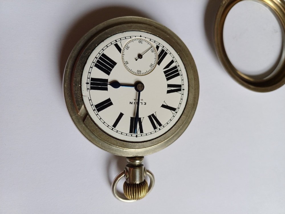An open-face antique pocket watch featuring a white enamel dial with black Roman numerals, a sub-seconds dial, and a silver-tone nickel case. View IV