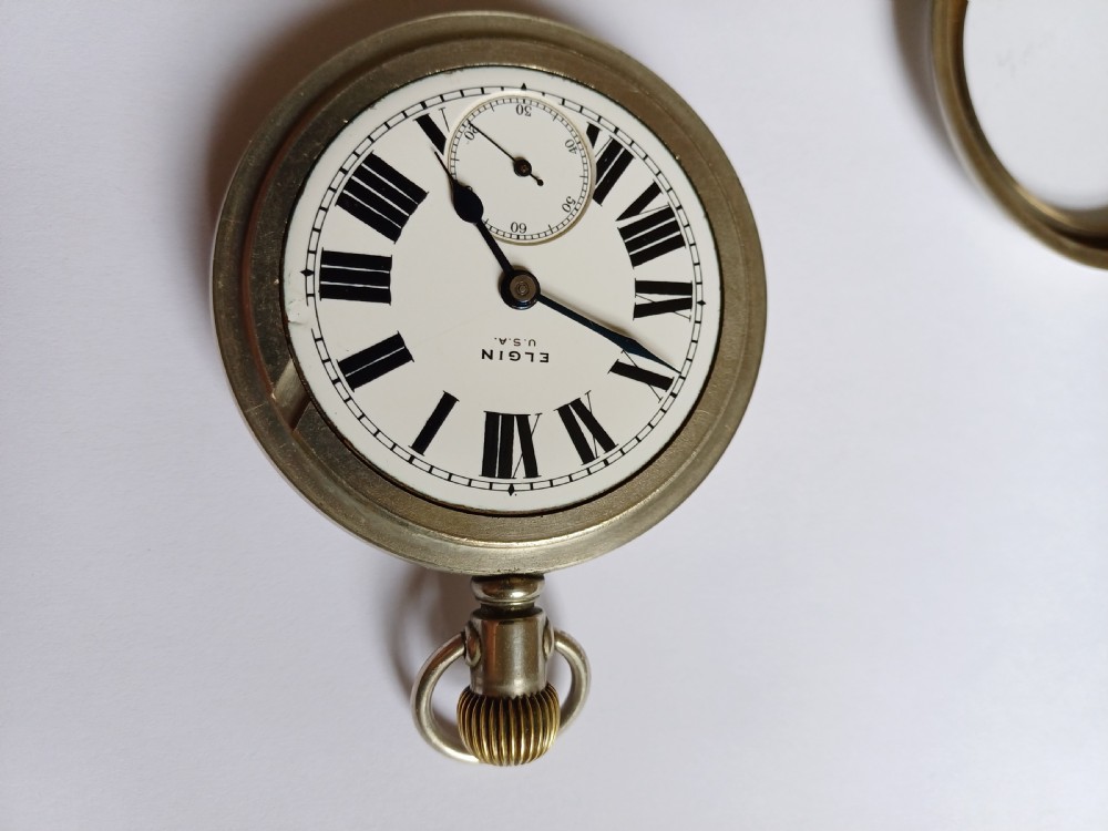 An open-face antique pocket watch featuring a white enamel dial with black Roman numerals, a sub-seconds dial, and a silver-tone nickel case. View V