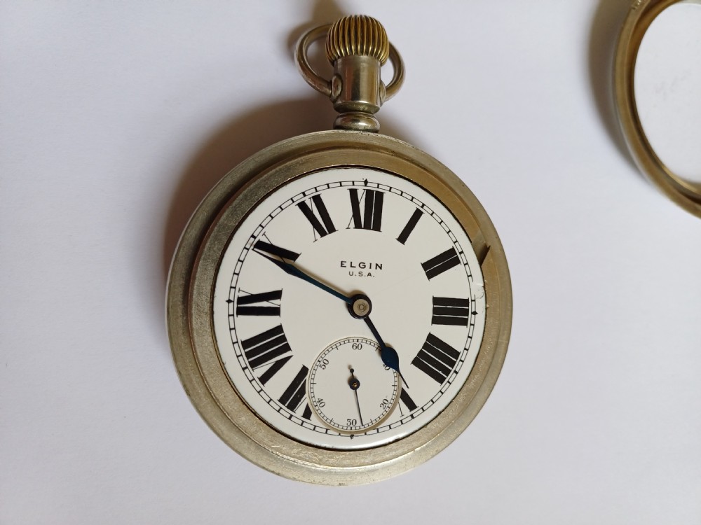 An open-face antique pocket watch featuring a white enamel dial with black Roman numerals, a sub-seconds dial, and a silver-tone nickel case. View VI