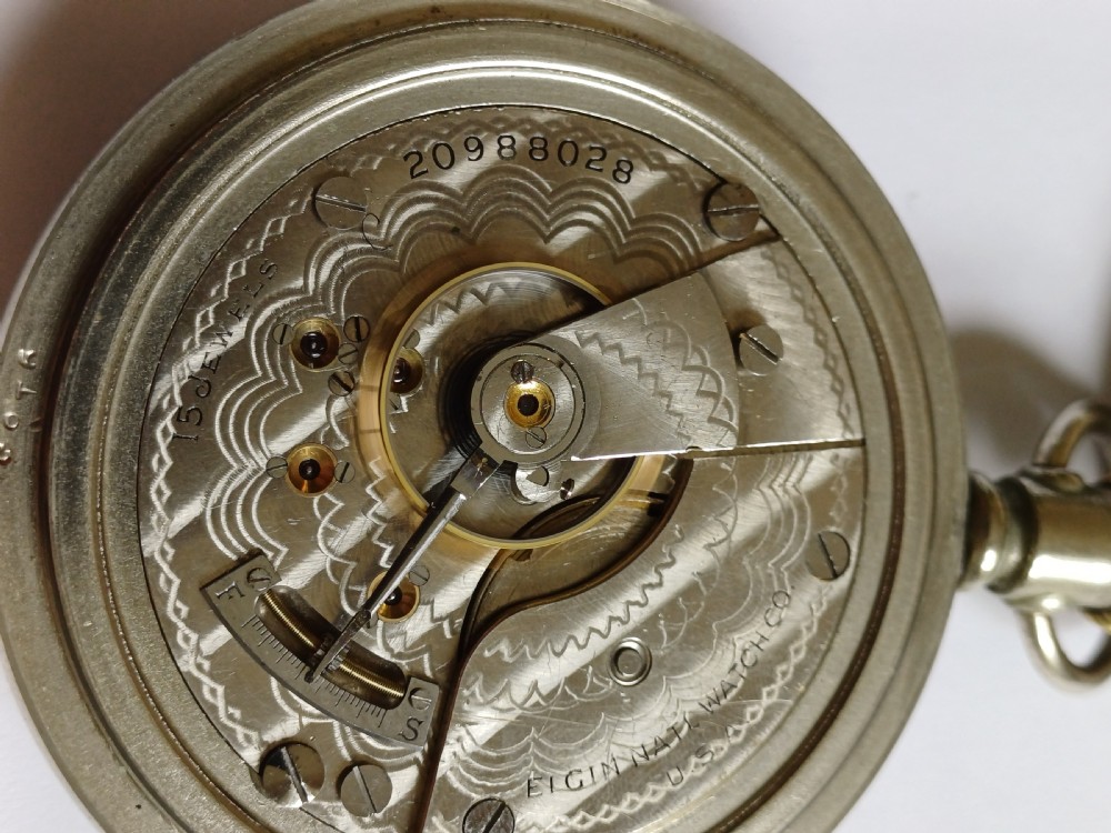 An open-face antique pocket watch featuring a white enamel dial with black Roman numerals, a sub-seconds dial, and a silver-tone nickel case. View XII