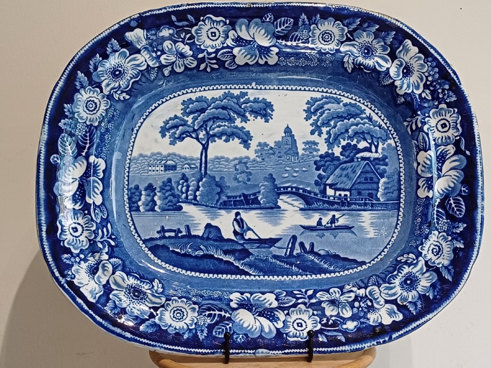 A large Victorian oval earthenware platter decorated with a cobalt blue and white transfer print featuring a pastoral river scene and a wide floral border.