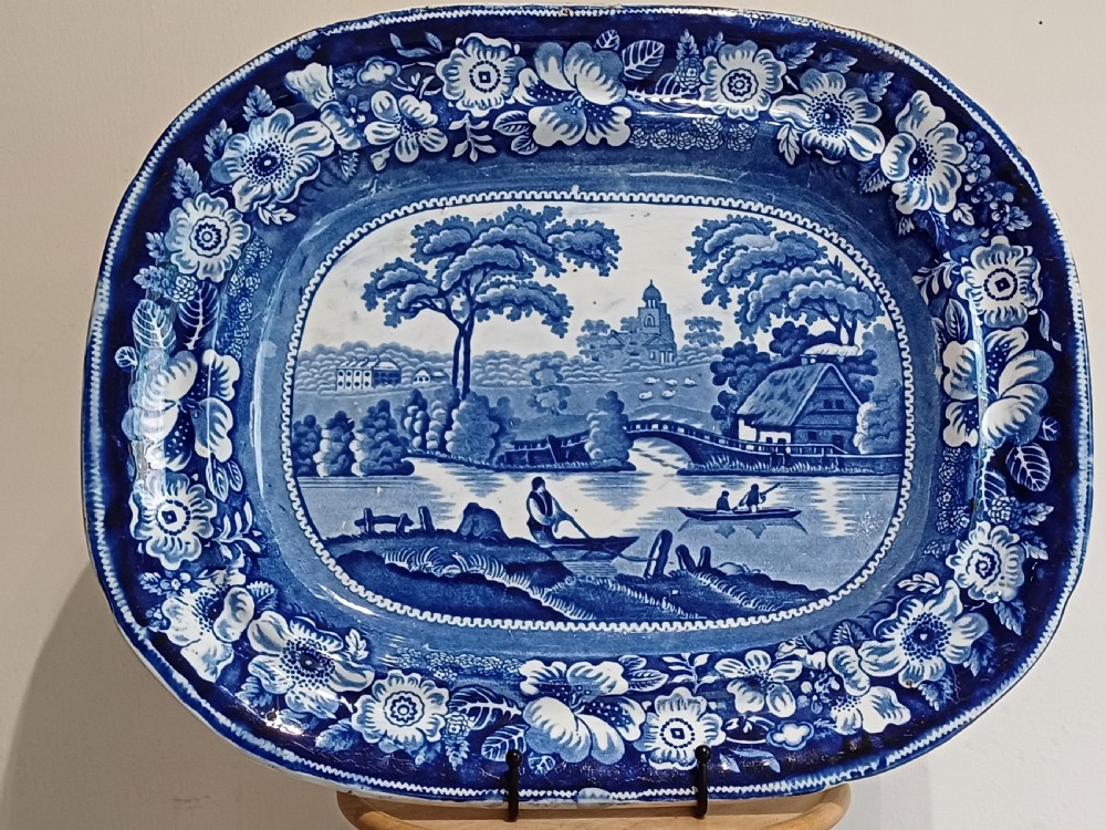 A large Victorian oval earthenware platter decorated with a cobalt blue and white transfer print featuring a pastoral river scene and a wide floral border. View II
