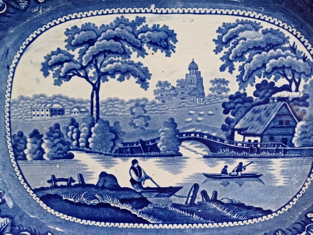 A large Victorian oval earthenware platter decorated with a cobalt blue and white transfer print featuring a pastoral river scene and a wide floral border. View V