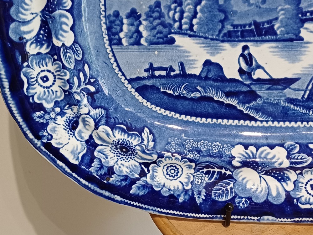 A large Victorian oval earthenware platter decorated with a cobalt blue and white transfer print featuring a pastoral river scene and a wide floral border. View VI
