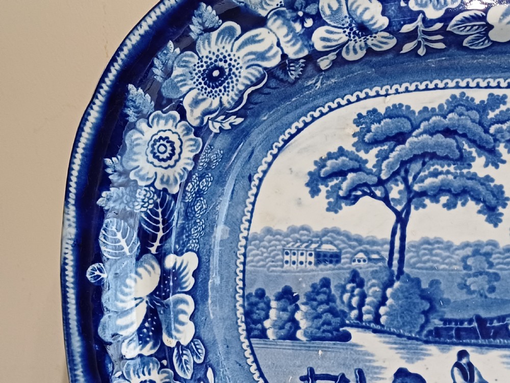 A large Victorian oval earthenware platter decorated with a cobalt blue and white transfer print featuring a pastoral river scene and a wide floral border. View VII