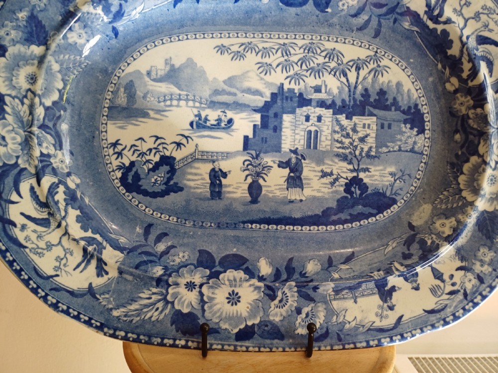 A large oval blue and white earthenware meat platter featuring a transfer-printed Chinese garden scene with figures, bridges, and floral borders. View V