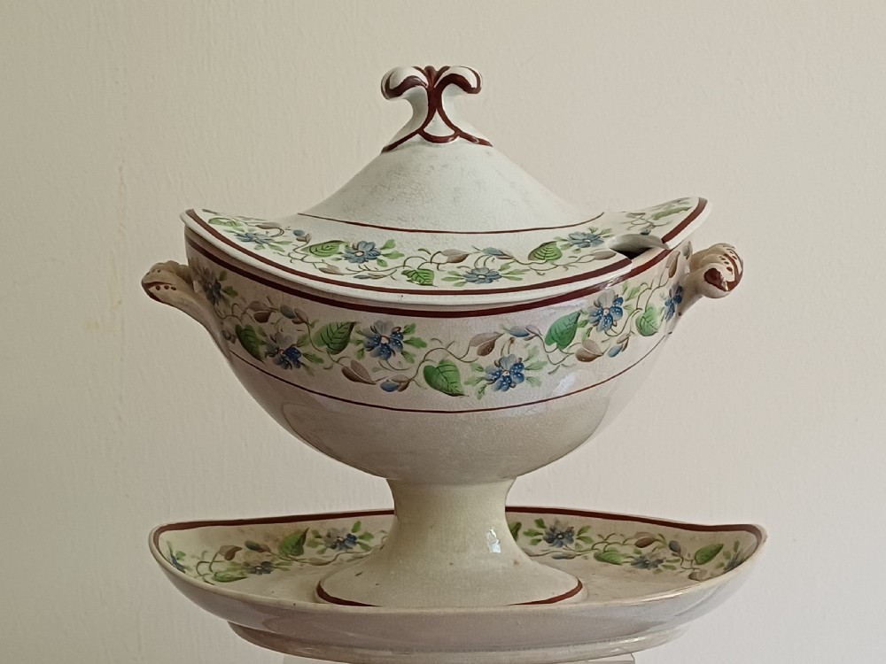 An antique cream earthenware pedestal dessert tureen with matching lid and stand, decorated with a floral vine border in blue and green.