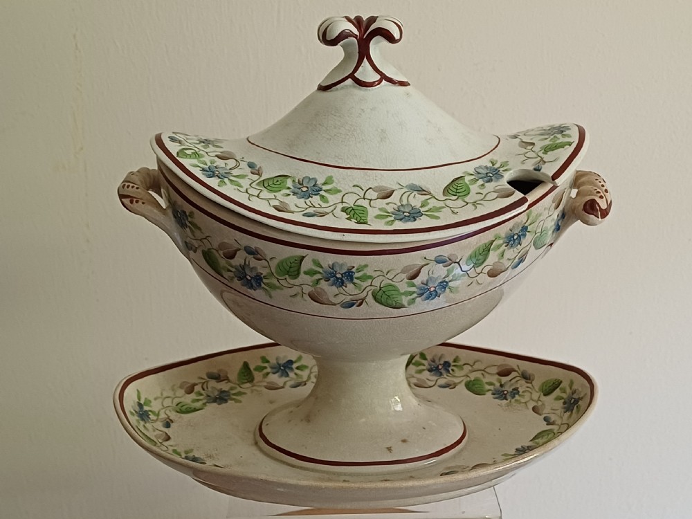 An antique cream earthenware pedestal dessert tureen with matching lid and stand, decorated with a floral vine border in blue and green. View XX