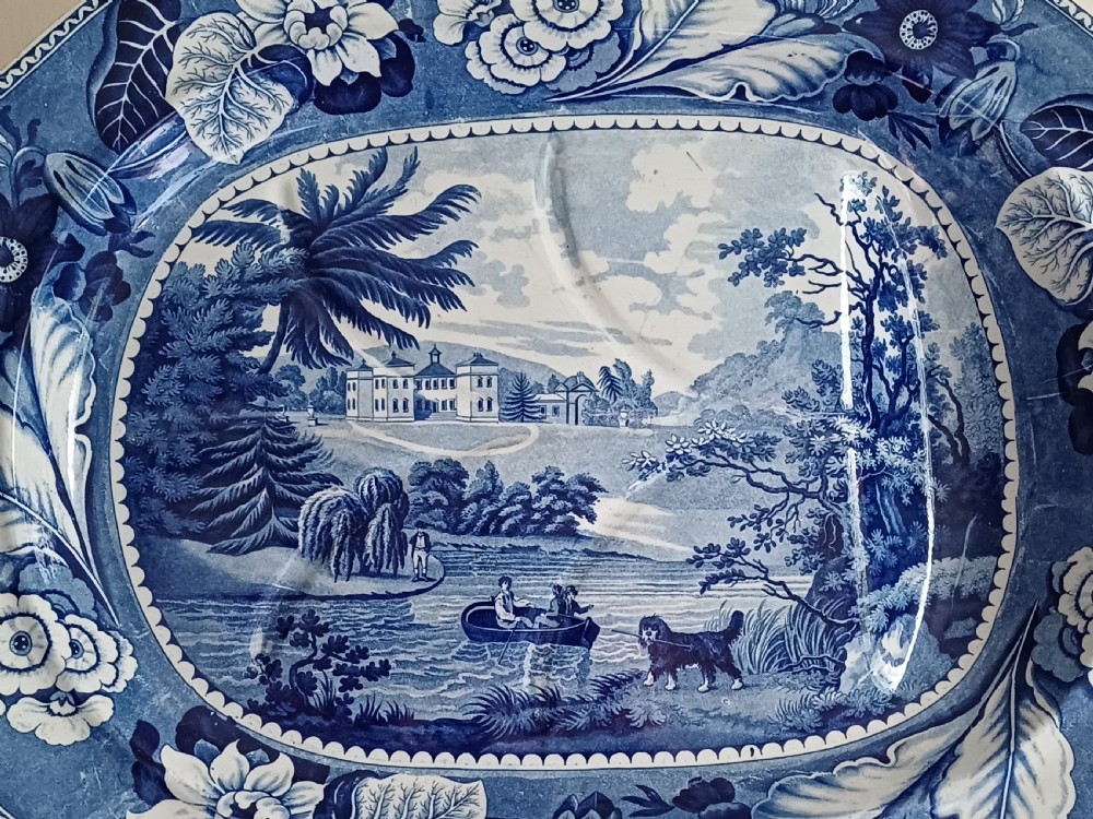 A large oval blue and white transfer printed earthenware meat platter featuring a country landscape scene with a dog and a moulded gravy well channel. View II