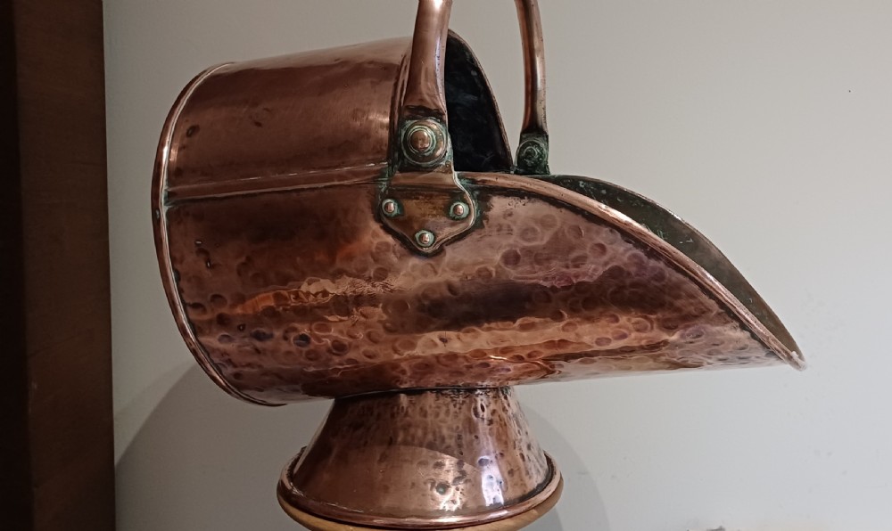 A hand-hammered copper helmet-shaped coal scuttle featuring a curved handle, riveted mountings, and a flared pedestal base.