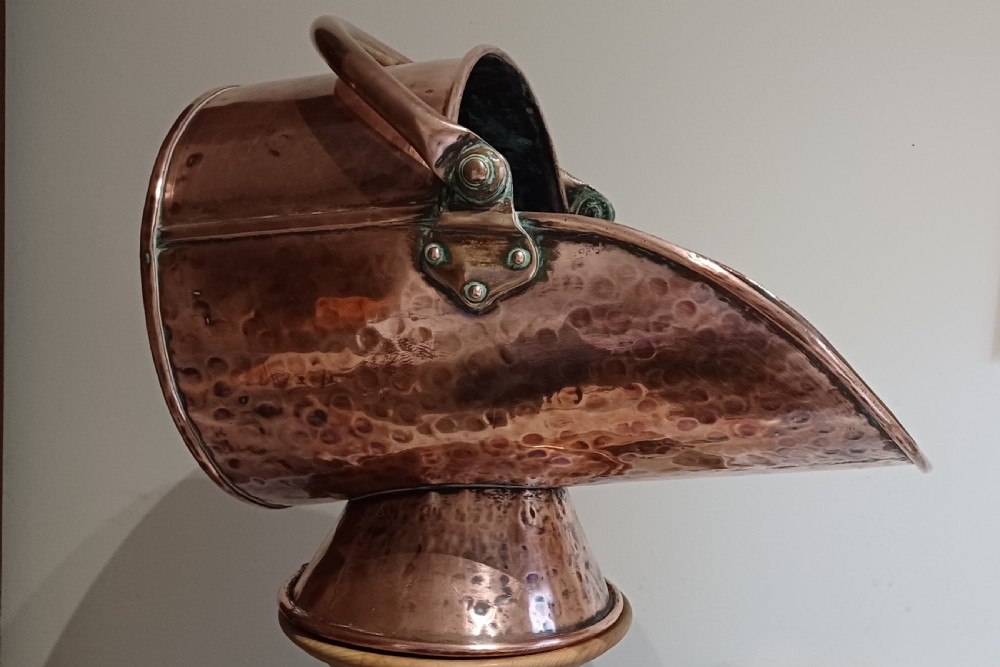 A hand-hammered copper helmet-shaped coal scuttle featuring a curved handle, riveted mountings, and a flared pedestal base. View VIII