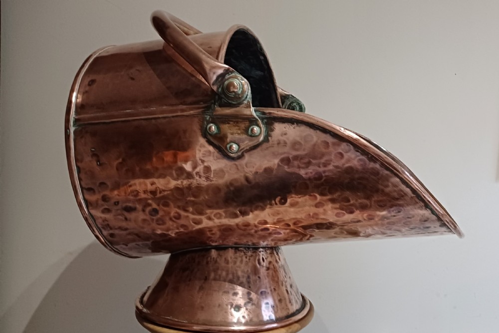 A hand-hammered copper helmet-shaped coal scuttle featuring a curved handle, riveted mountings, and a flared pedestal base. View XII