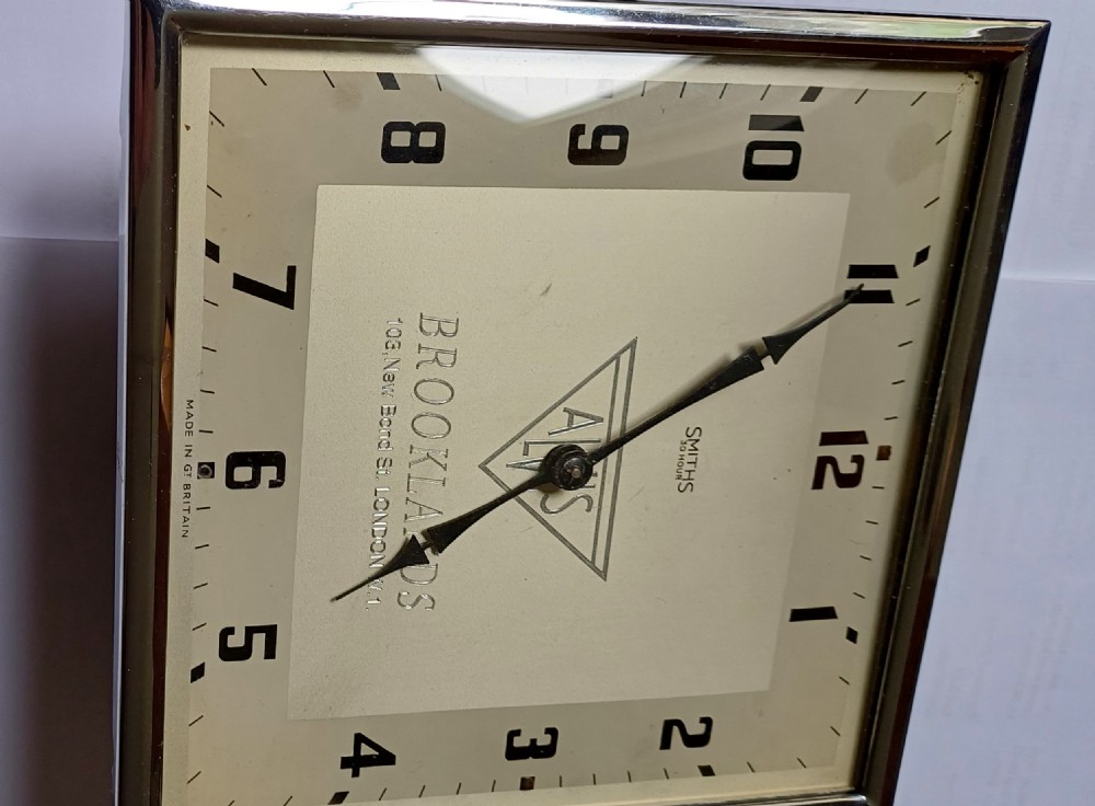 A square mid-century chrome desk clock featuring a cream dial with black Arabic numerals and embossed Alvis and Brooklands branding. View VIII
