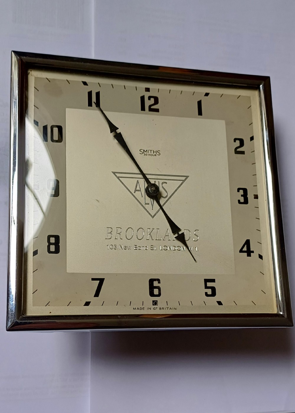 A square mid-century chrome desk clock featuring a cream dial with black Arabic numerals and embossed Alvis and Brooklands branding. View XI