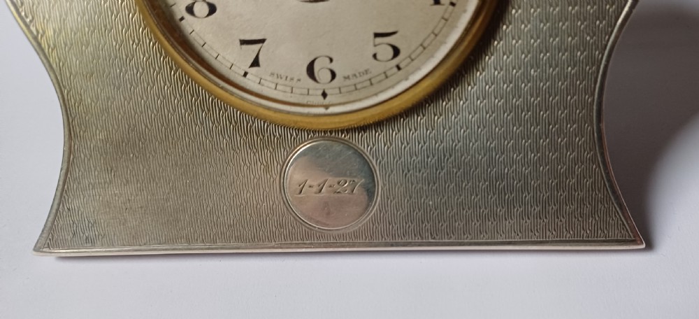 An Art Deco engine-turned silver strut desk clock featuring a shaped outline, circular dial with Arabic numerals, and an engraved circular plaque dated 1927. View IV