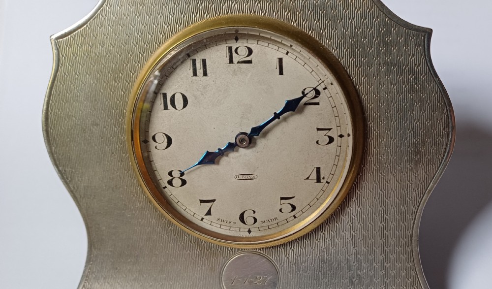 An Art Deco engine-turned silver strut desk clock featuring a shaped outline, circular dial with Arabic numerals, and an engraved circular plaque dated 1927. View V