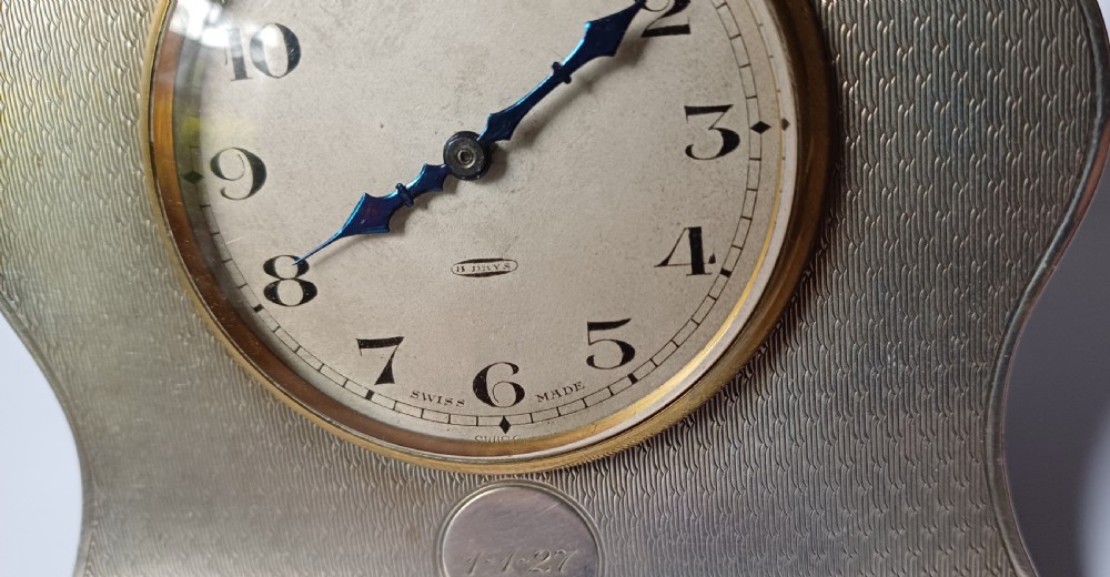 An Art Deco engine-turned silver strut desk clock featuring a shaped outline, circular dial with Arabic numerals, and an engraved circular plaque dated 1927. View VI