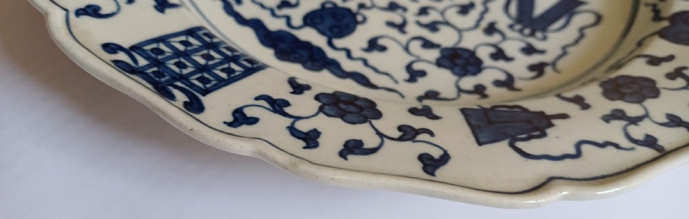 An 18th-century circular soft-paste porcelain plate with a scalloped rim, featuring underglaze cobalt blue Chinese-inspired motifs and scrolling foliage. View VIII