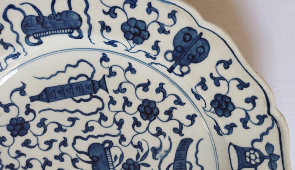 An 18th-century circular soft-paste porcelain plate with a scalloped rim, featuring underglaze cobalt blue Chinese-inspired motifs and scrolling foliage. View X