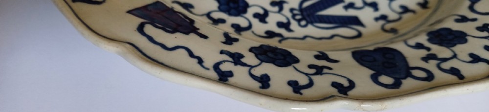 An 18th-century circular soft-paste porcelain plate with a scalloped rim, featuring underglaze cobalt blue Chinese-inspired motifs and scrolling foliage. View XIV