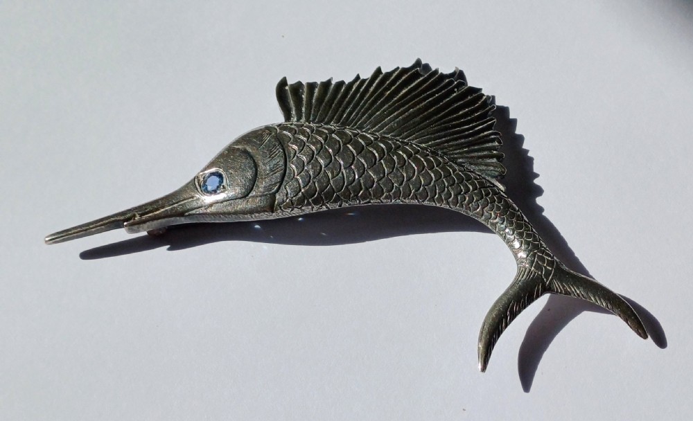 A vintage silver brooch shaped as a sailfish with detailed scales, a prominent ribbed dorsal fin, and a light blue stone set in the eye.