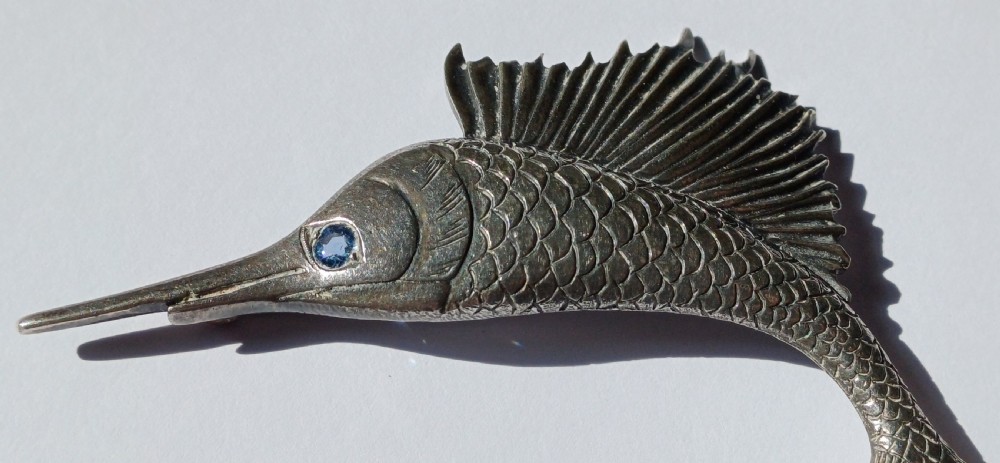 A vintage silver brooch shaped as a sailfish with detailed scales, a prominent ribbed dorsal fin, and a light blue stone set in the eye. View II