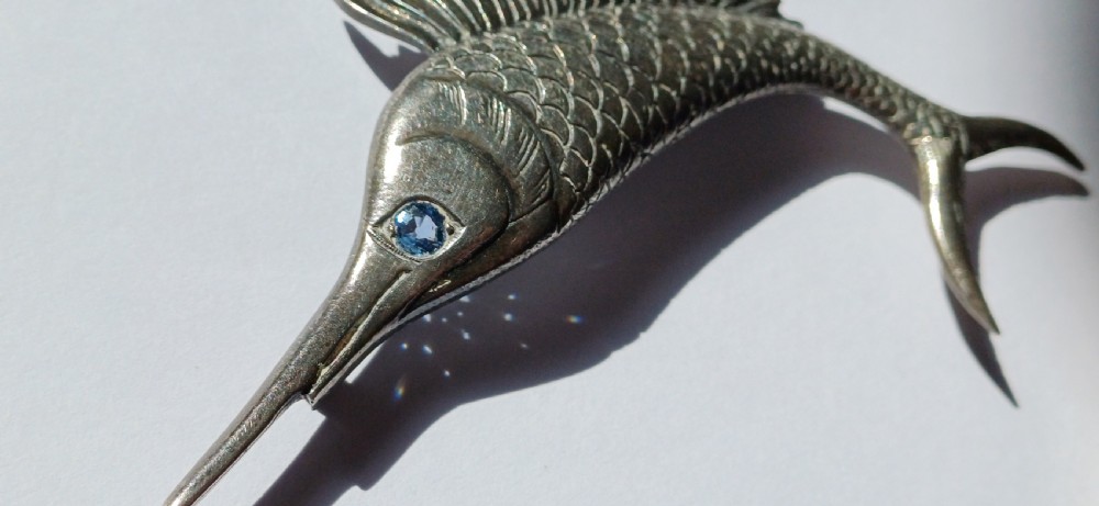 A vintage silver brooch shaped as a sailfish with detailed scales, a prominent ribbed dorsal fin, and a light blue stone set in the eye. View VI