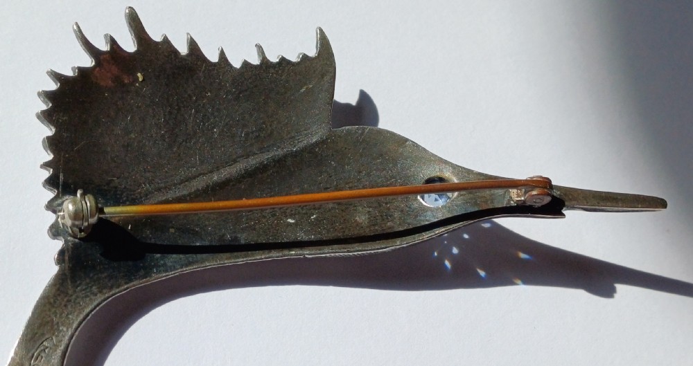 A vintage silver brooch shaped as a sailfish with detailed scales, a prominent ribbed dorsal fin, and a light blue stone set in the eye. View IX