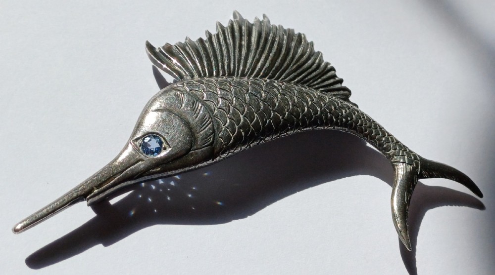 A vintage silver brooch shaped as a sailfish with detailed scales, a prominent ribbed dorsal fin, and a light blue stone set in the eye. View XI