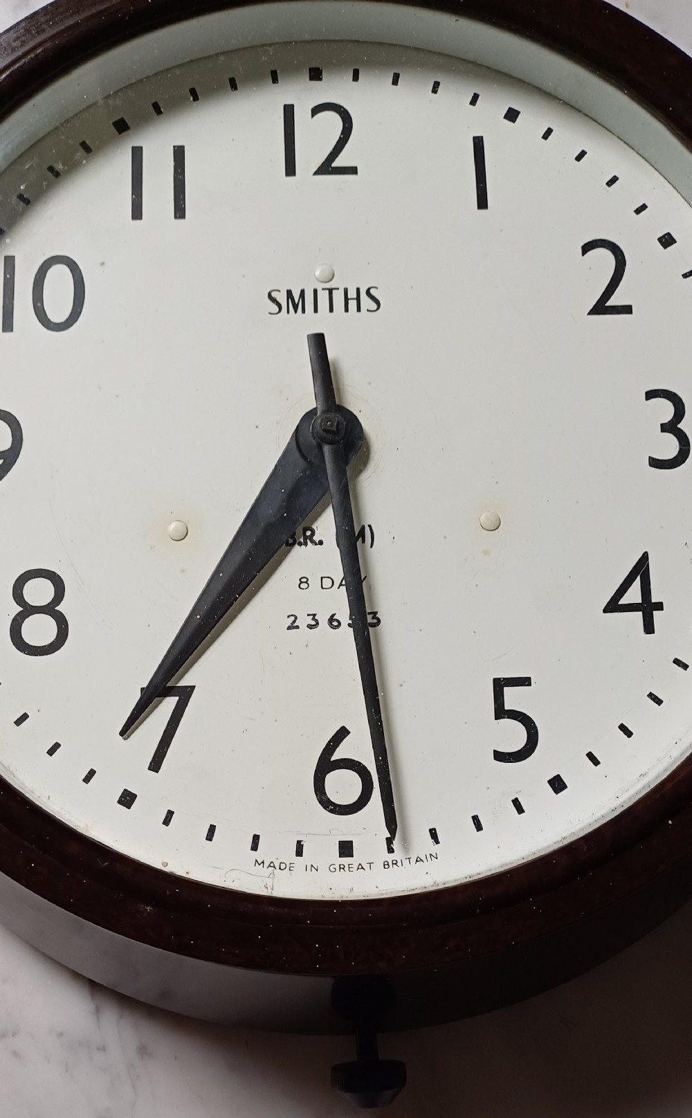 A circular mid-century wall clock featuring a moulded dark brown Bakelite case, a white dial with black Arabic numerals, and dark hands. View II