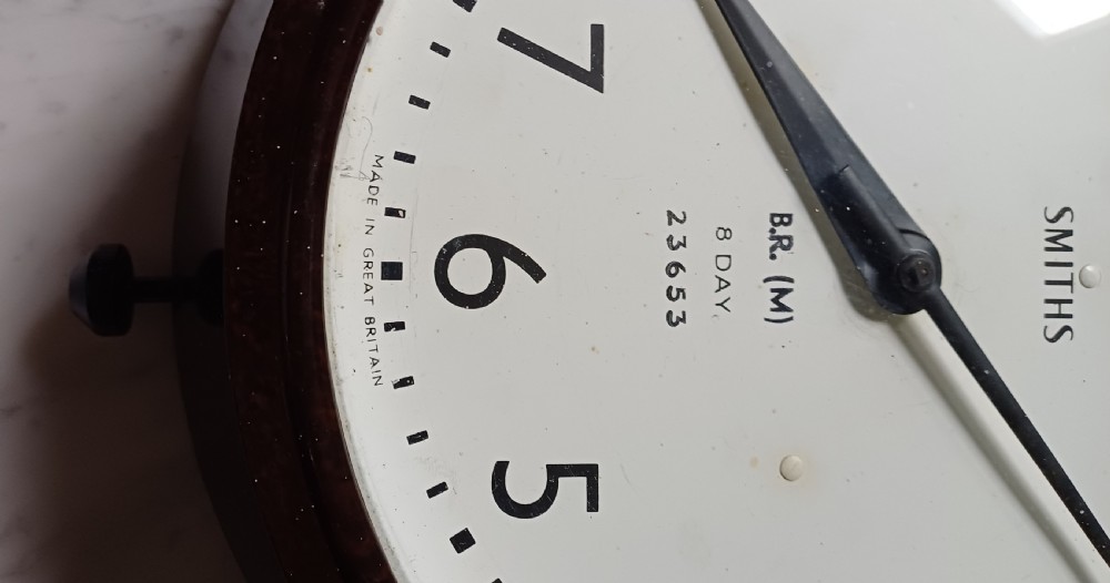 A circular mid-century wall clock featuring a moulded dark brown Bakelite case, a white dial with black Arabic numerals, and dark hands. View V