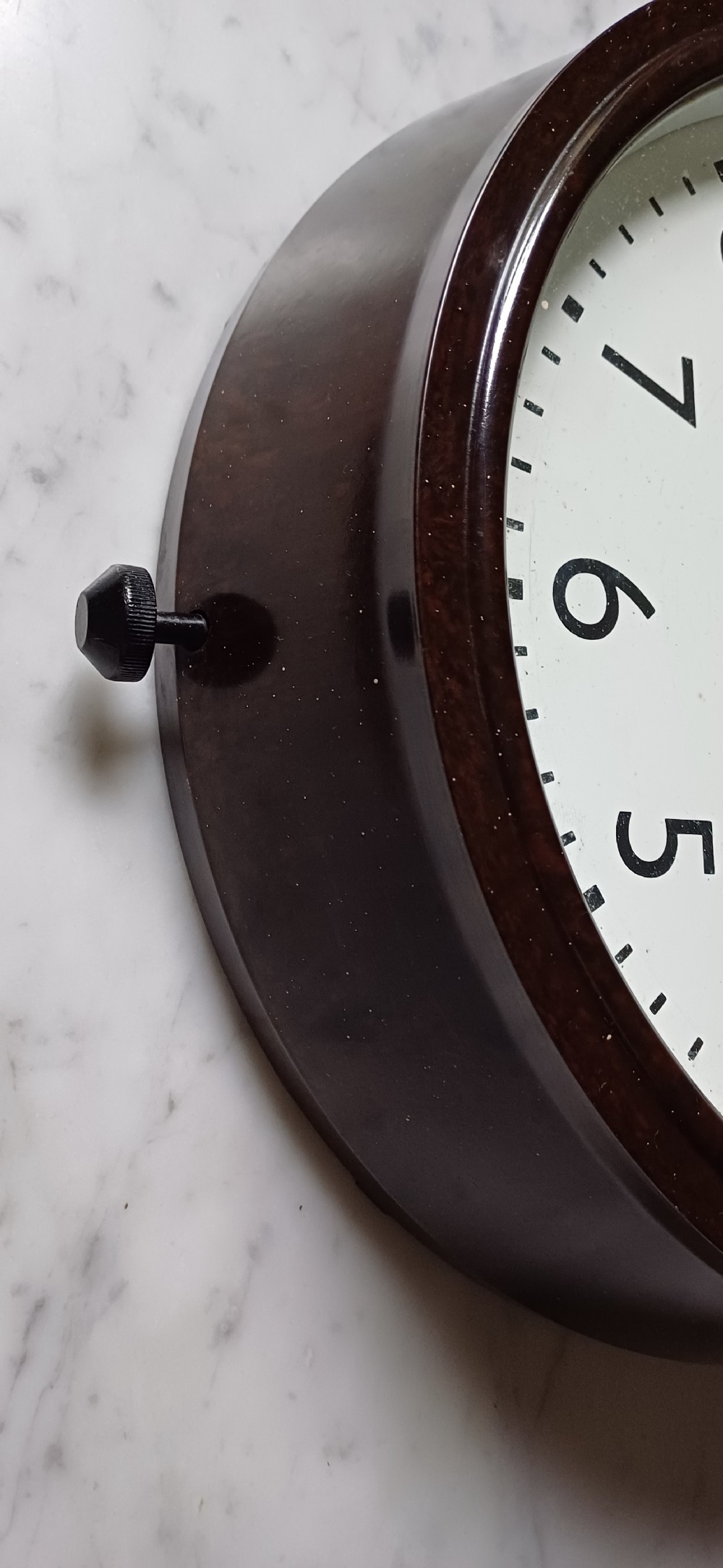 A circular mid-century wall clock featuring a moulded dark brown Bakelite case, a white dial with black Arabic numerals, and dark hands. View VII