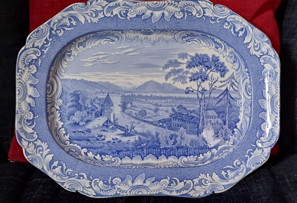 A large oval blue and white earthenware serving platter featuring an underglaze transfer-printed landscape scene framed by an ornate scroll border.