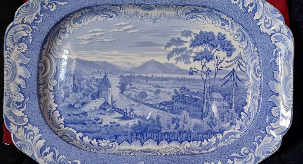 A large oval blue and white earthenware serving platter featuring an underglaze transfer-printed landscape scene framed by an ornate scroll border. View II