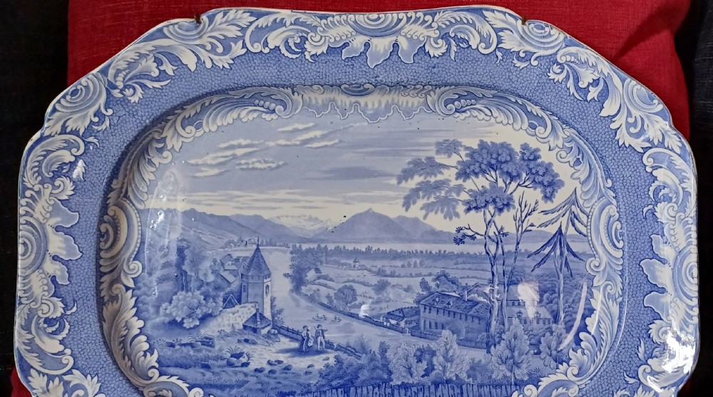 A large oval blue and white earthenware serving platter featuring an underglaze transfer-printed landscape scene framed by an ornate scroll border. View IV