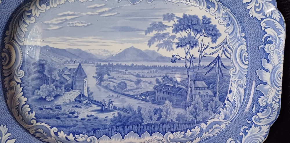 A large oval blue and white earthenware serving platter featuring an underglaze transfer-printed landscape scene framed by an ornate scroll border. View V