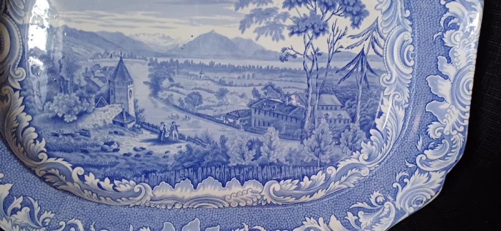 A large oval blue and white earthenware serving platter featuring an underglaze transfer-printed landscape scene framed by an ornate scroll border. View VI