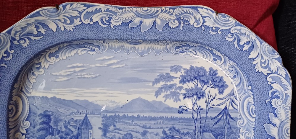A large oval blue and white earthenware serving platter featuring an underglaze transfer-printed landscape scene framed by an ornate scroll border. View VII