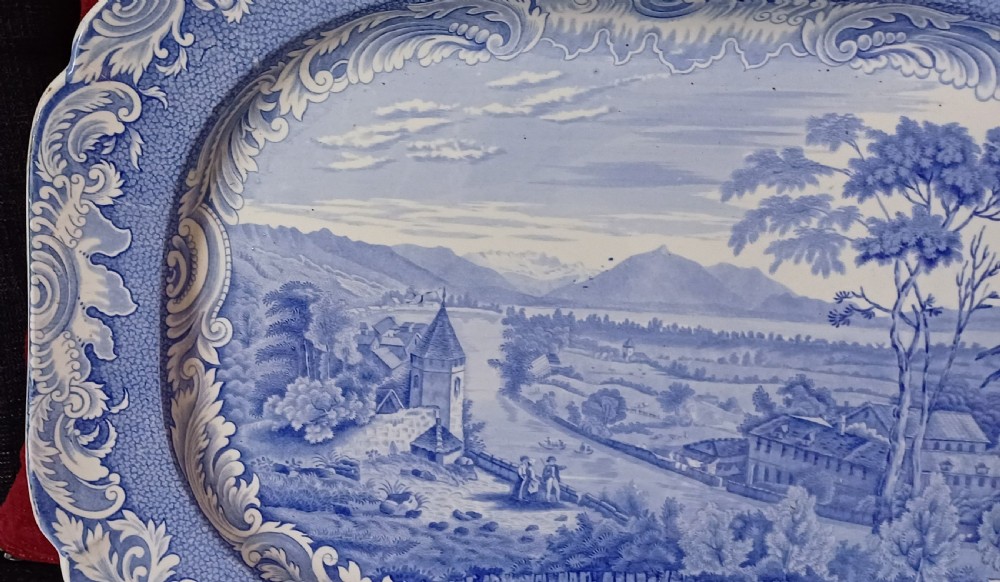 A large oval blue and white earthenware serving platter featuring an underglaze transfer-printed landscape scene framed by an ornate scroll border. View IX