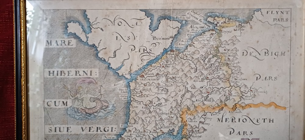 A framed 17th-century hand-coloured map of Caernarvonshire featuring a decorative cartouche, compass rose, and sea monster illustration. View IX