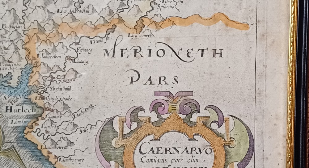 A framed 17th-century hand-coloured map of Caernarvonshire featuring a decorative cartouche, compass rose, and sea monster illustration. View X