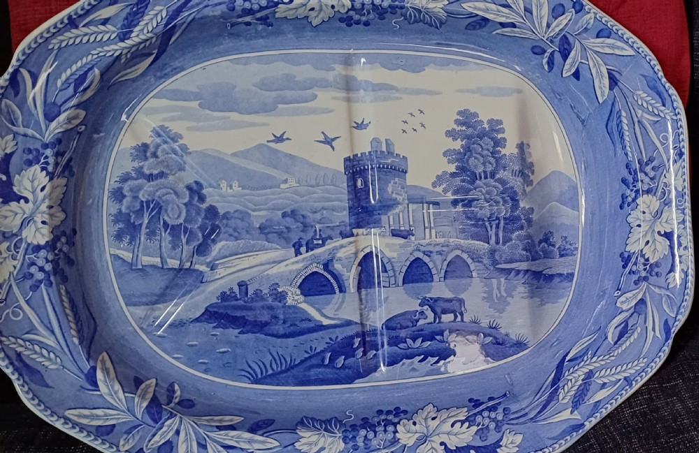 An oval blue and white transfer-printed earthenware meat platter depicting a central landscape with a bridge, tower, and cattle framed by a broad vine leaf border. View III