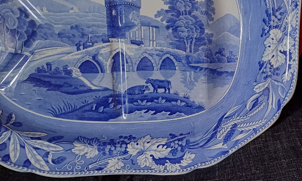 An oval blue and white transfer-printed earthenware meat platter depicting a central landscape with a bridge, tower, and cattle framed by a broad vine leaf border. View IV