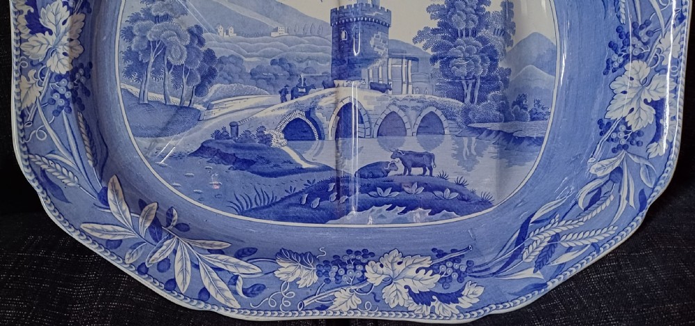 An oval blue and white transfer-printed earthenware meat platter depicting a central landscape with a bridge, tower, and cattle framed by a broad vine leaf border. View V